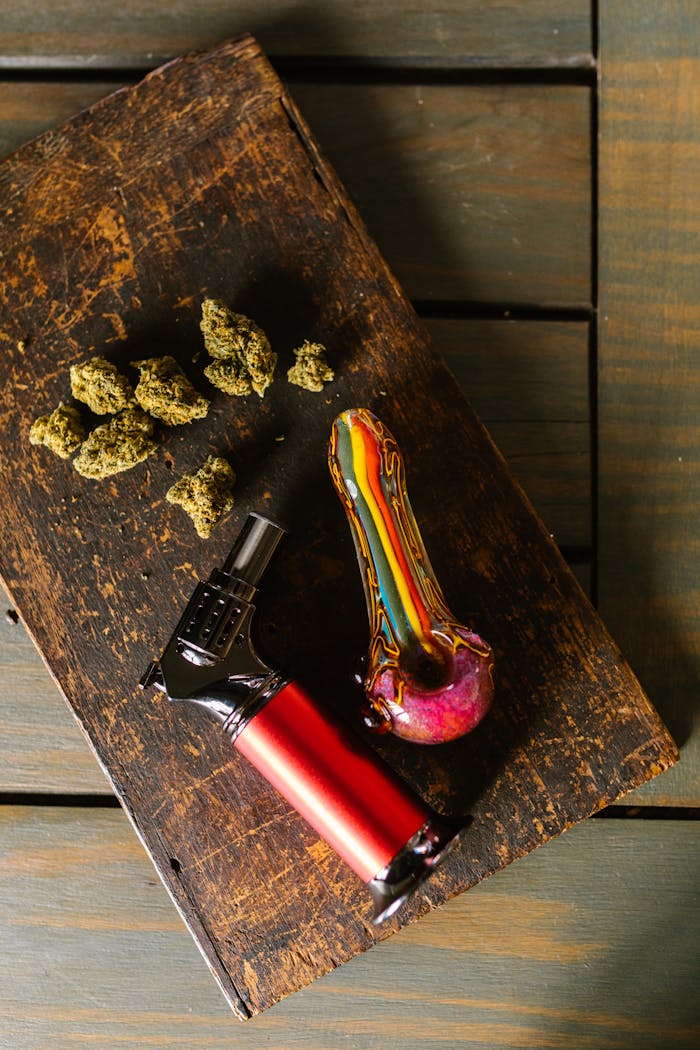 Top view of cannabis buds, colorful glass pipe, and lighter on rustic tray.