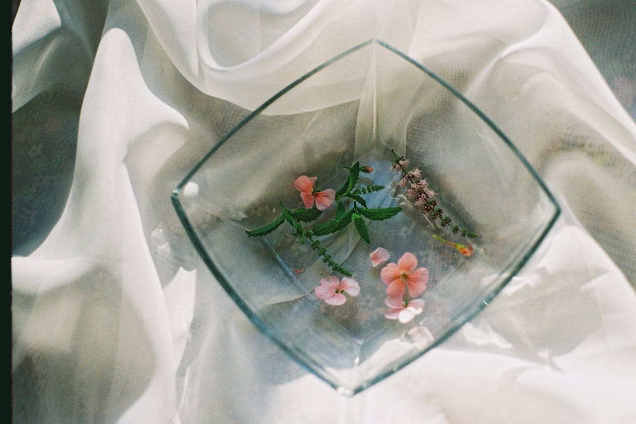 Square glass bowl with floating pink flowers and leaves on white fabric. Elegant and serene composition.